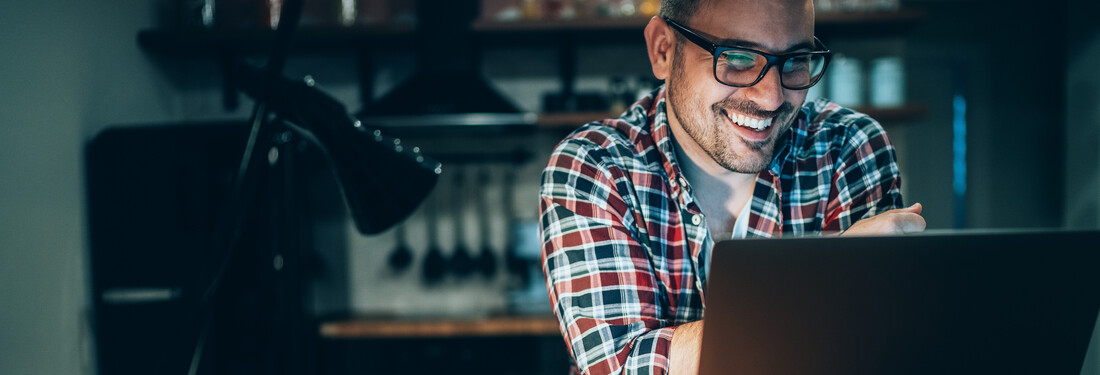 A man with glasses smiles while using his laptop, reflecting a positive and focused work environment.